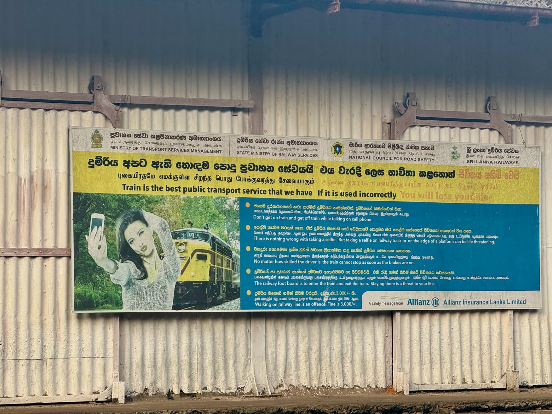 Sri Lanka Railways safety sign on a corrugated wall, with a woman taking a selfie beside a train